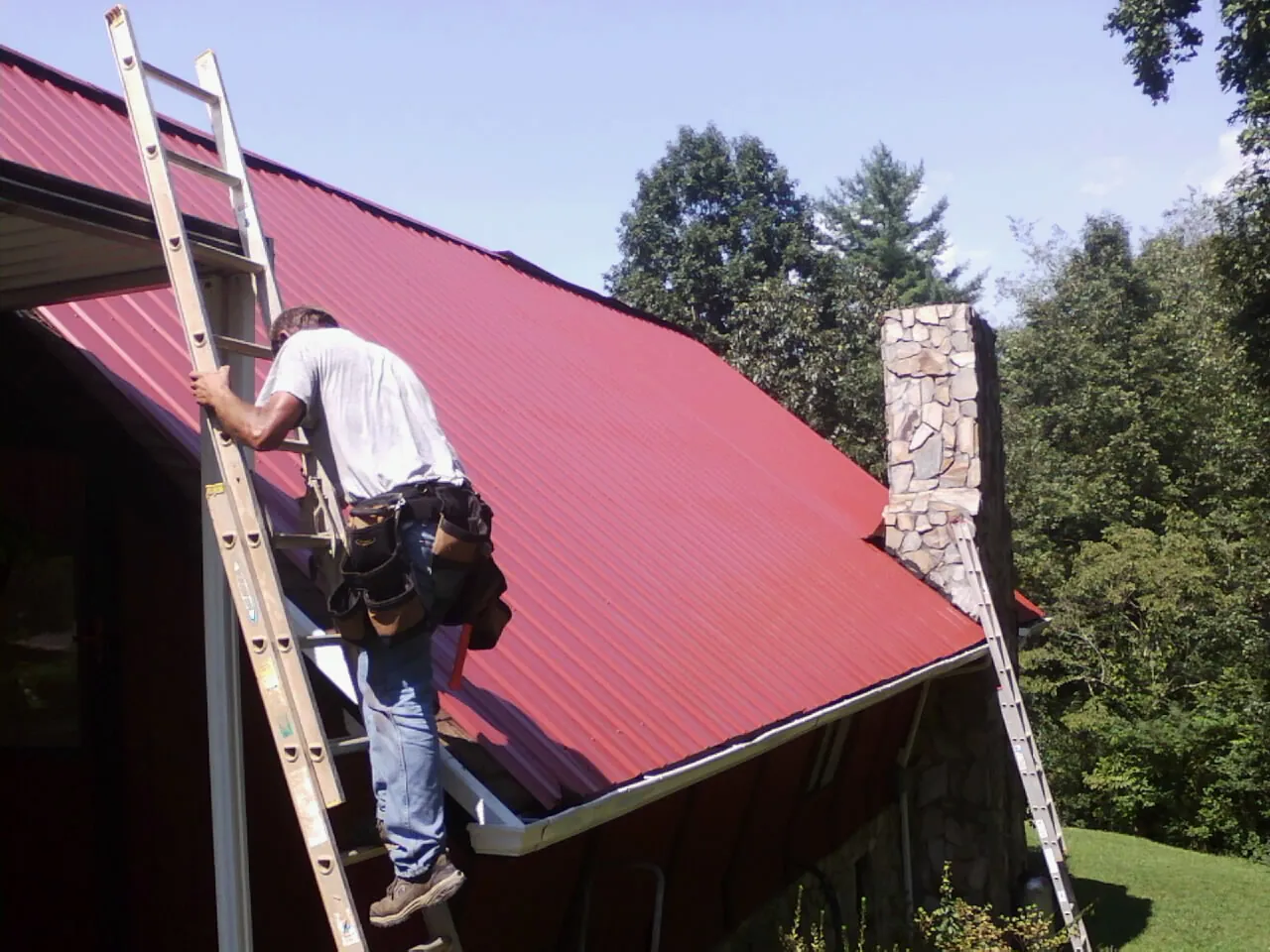 Roofer on a ladder beside a red metal roof and stone chimney — Soffit & Fascia Repair by Roofing Brothers in Joseph